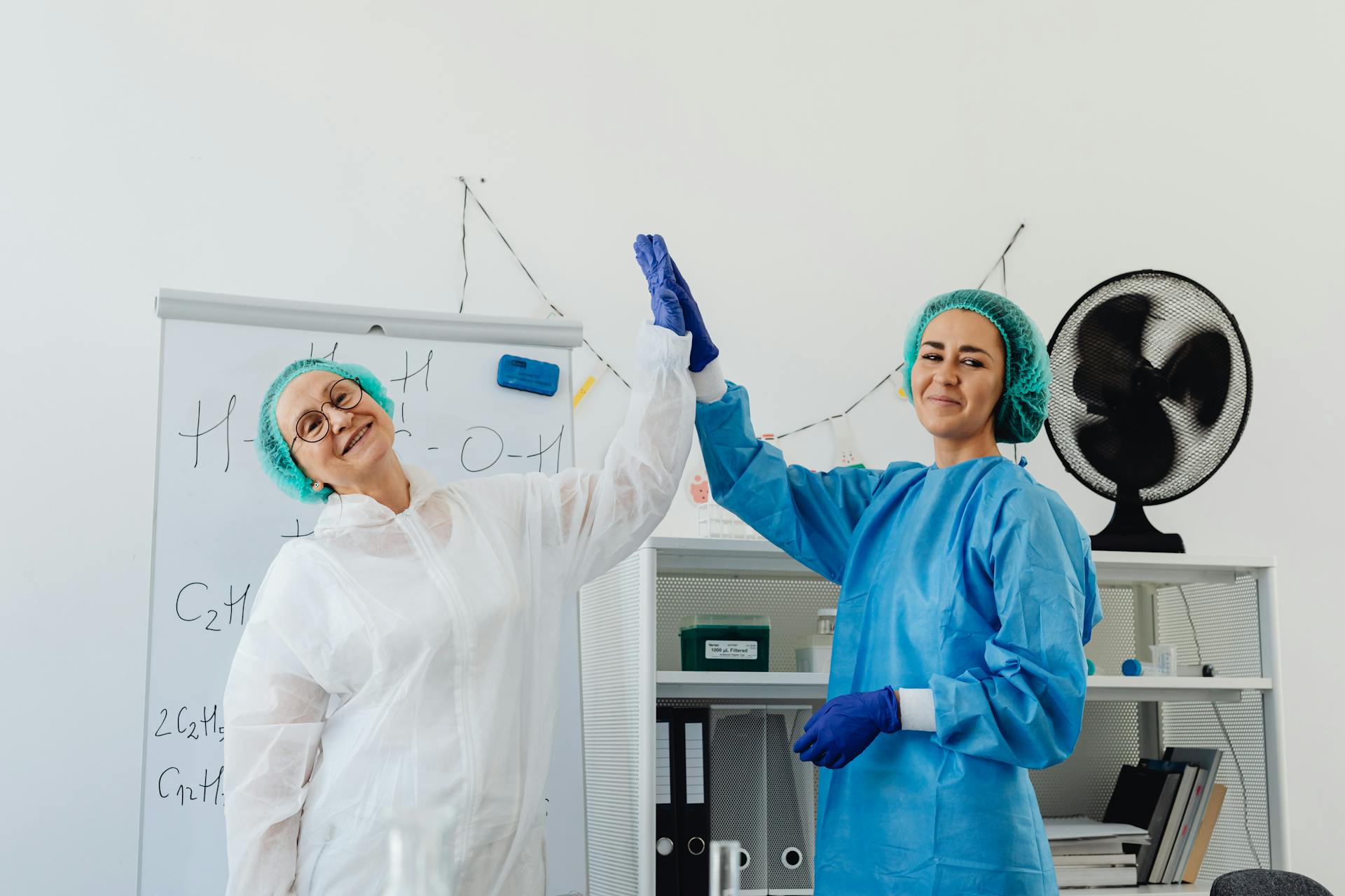 Two female healthcare professionals in protective outfits high-fiving in a lab.