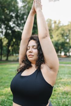 Woman practicing yoga with eyes closed, arms raised in serene park setting, promoting mindfulness and wellness.