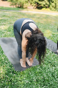 Woman performing yoga stretch on mat in sunny park setting.