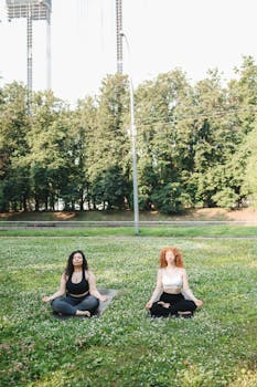 Two women practicing yoga meditation in a sunny park, promoting relaxation and wellness.