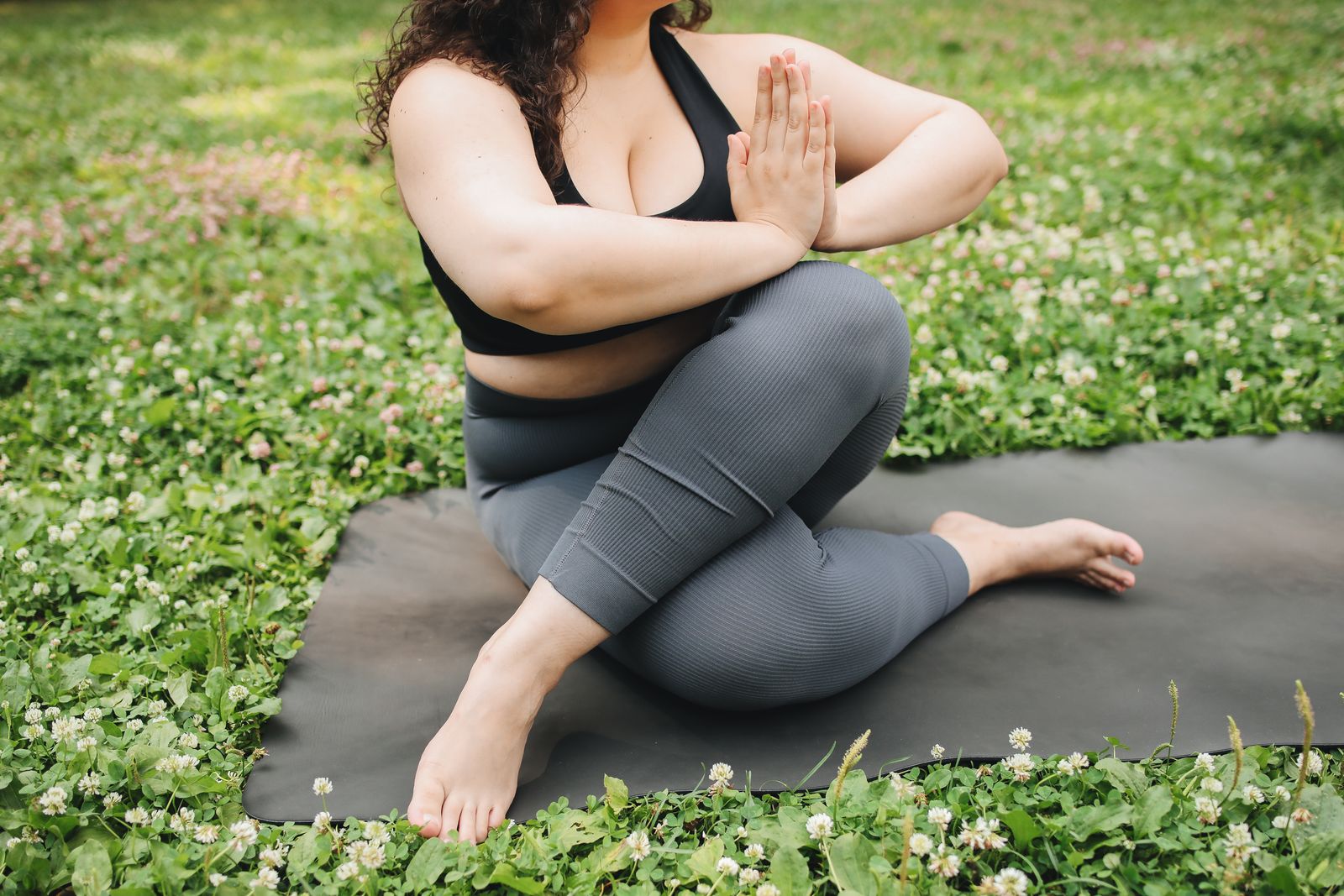 Plus-size woman practicing a standing yoga pose outdoors on a mat surrounded by greenery