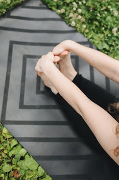 A woman performing a stretching exercise on a black yoga mat in a lush green outdoor setting.
