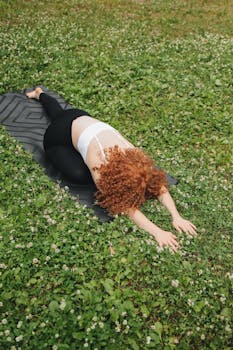 Curly-haired woman doing yoga stretches on a mat in a grassy park.