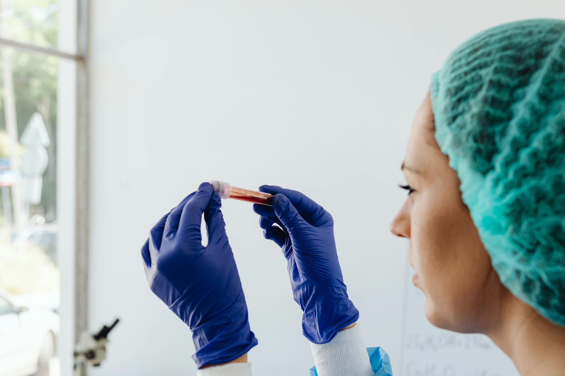 A focused lab technician holds a blood sample in a test tube, wearing gloves and a hair net.