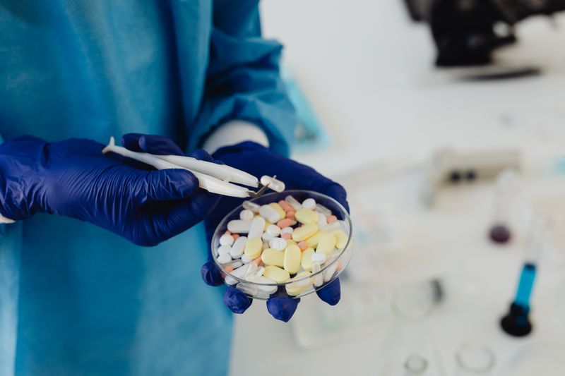 A researcher in a blue surgical gown and blue nitrile gloves uses tweezers to handle assorted prescription tablets in a petri dish, with a soft-focus lab bench in the background.