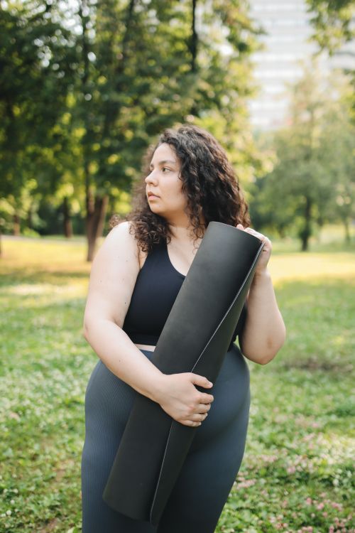 Free Plus-sized woman in activewear holding a yoga mat outdoors in a sunny park setting. Stock Photo