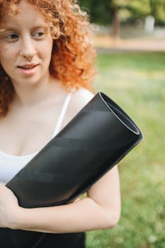 A young woman with curly hair holds a yoga mat outdoors on a sunny day, ready for a yoga session.