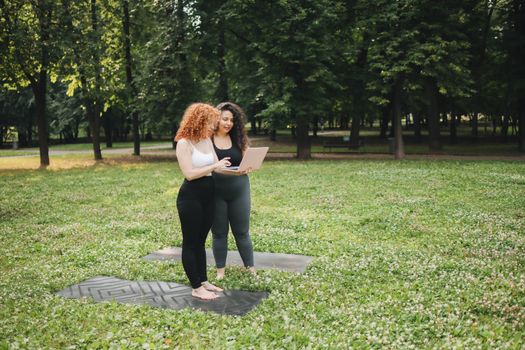 Two women on yoga mats using a laptop in a sunny park setting, surrounded by green grass and trees.