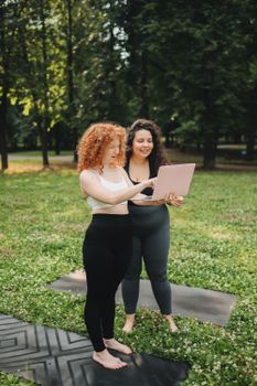 Two women in active wear enjoy a sunny day in the park while using a laptop on yoga mats.