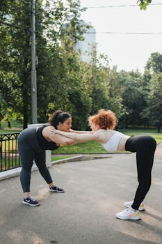 Two women in activewear stretching together in a sunny park setting.