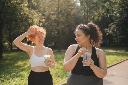 Free Two women in activewear enjoy refreshing drinks while relaxing outdoors in a sunny park. Stock Photo