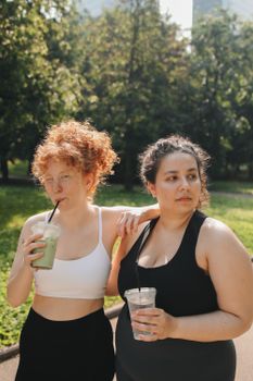 Two women in activewear enjoying drinks in a sunny park, promoting body positivity.