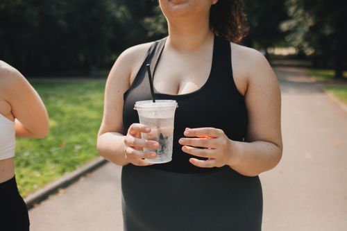 Free A plus size woman in a black tank top holding a drink while exercising in an outdoor park setting. Stock Photo