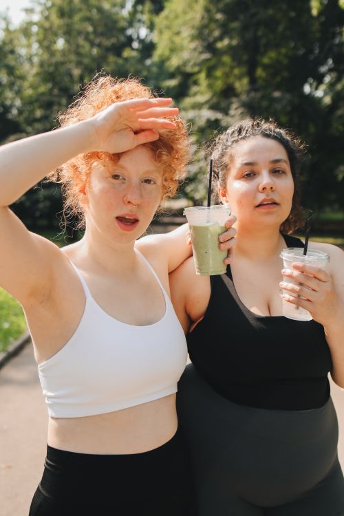 Free Two women in activewear enjoy refreshing drinks in a sunny park scene, promoting friendship and wellness. Stock Photo