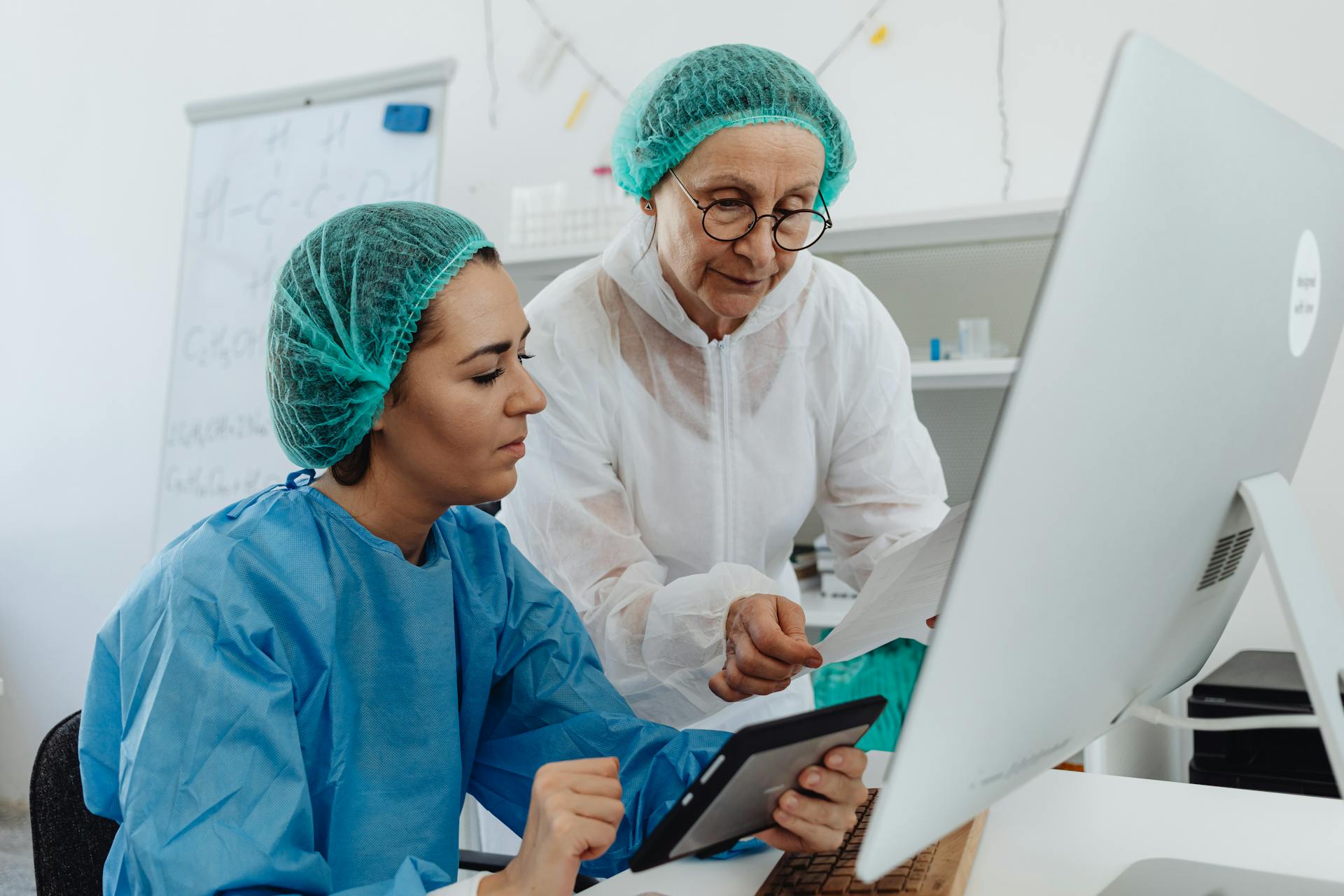 Two researchers in a laboratory examining data on a computer and tablet, wearing safety gear.