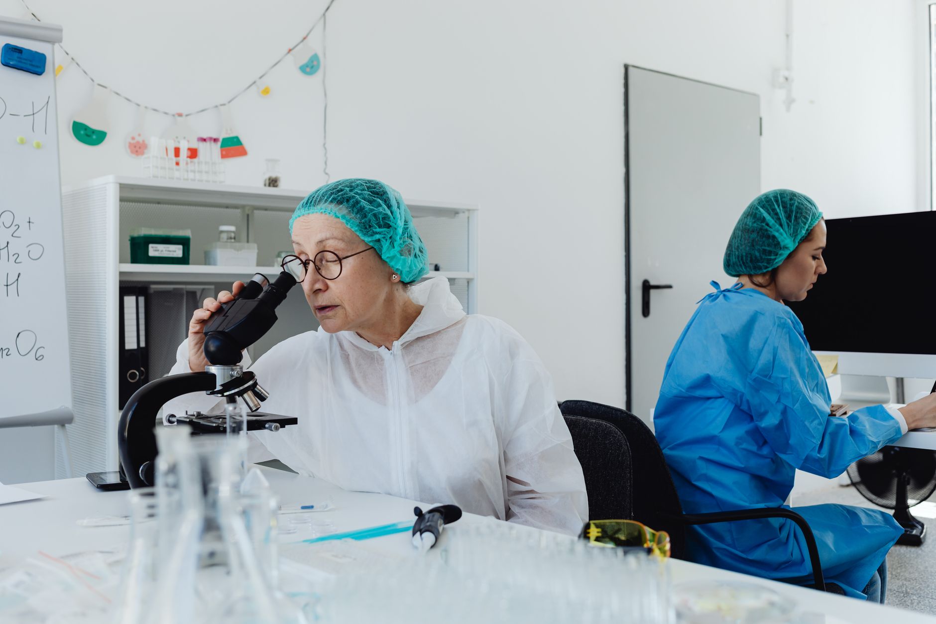 Scientists in coveralls and hair nets conducting research with microscopes and computers in a lab.