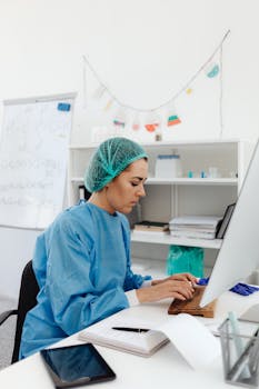 Medical professional in blue coveralls typing on a computer in a laboratory setting.