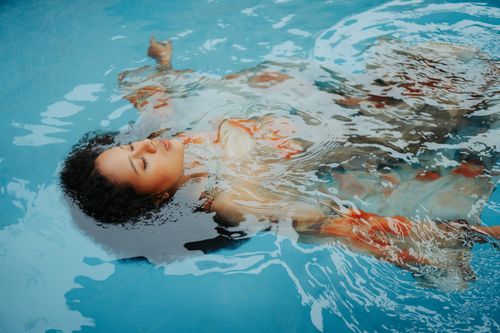 Free Serene woman in a flowing dress floating peacefully in a clear blue pool. Stock Photo