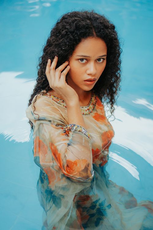Free A woman with curly hair in a floral dress gracefully poses in a pool, blending elegance with tranquility. Stock Photo
