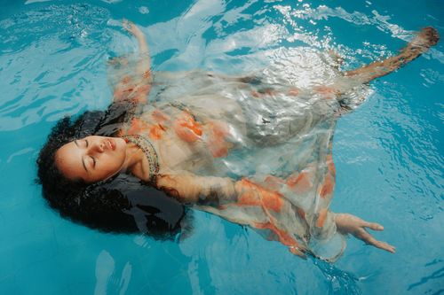 Free A woman in a floral dress floats peacefully with eyes closed in a clear blue swimming pool. Stock Photo