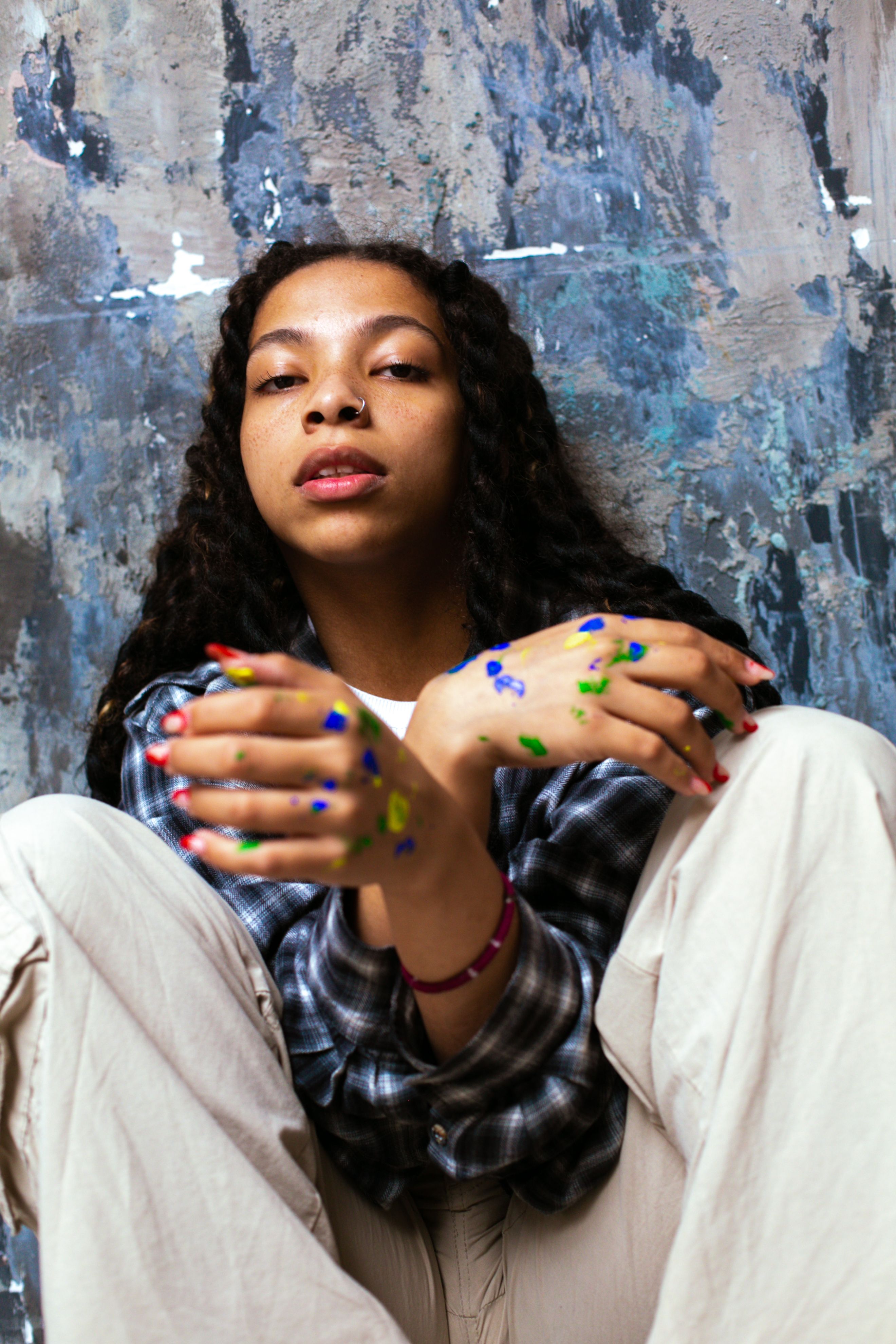 Young woman with paint-splattered hands sitting against a textured wall, expressing creativity and style.