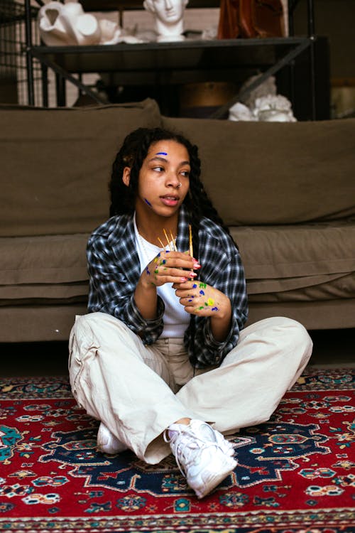 A Woman with Paint on Face and Hands Sitting on Floor Holding Paint Brushes