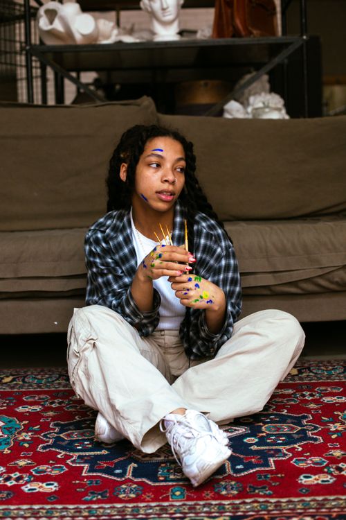 Free Young artist covered in paint holding brushes, sitting on colorful carpet in an art studio. Stock Photo