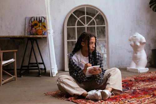 A Woman in Plaid Long Sleeves Sitting on a Rug while Writing on a Notebook