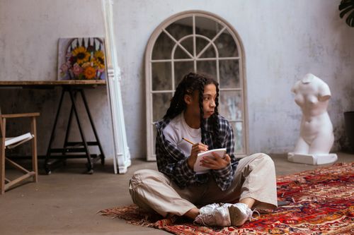 Free A creative woman sits on a patterned rug while sketching in an art studio with sculptures and paintings. Stock Photo