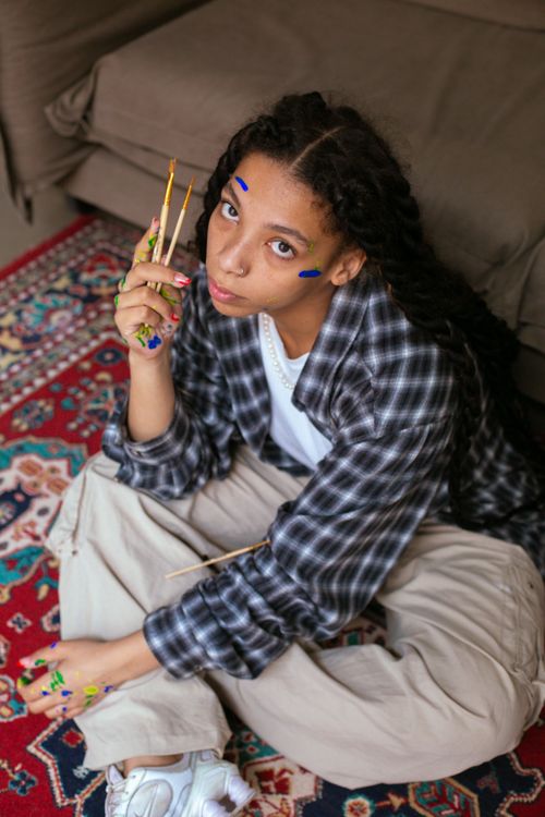 Free A young artist sitting indoors, holding paintbrushes with painted hands and face, exuding creativity. Stock Photo