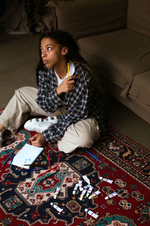 Free Young female artist sitting on a carpet with paints, contemplating her next creative project in a cozy setting. Stock Photo