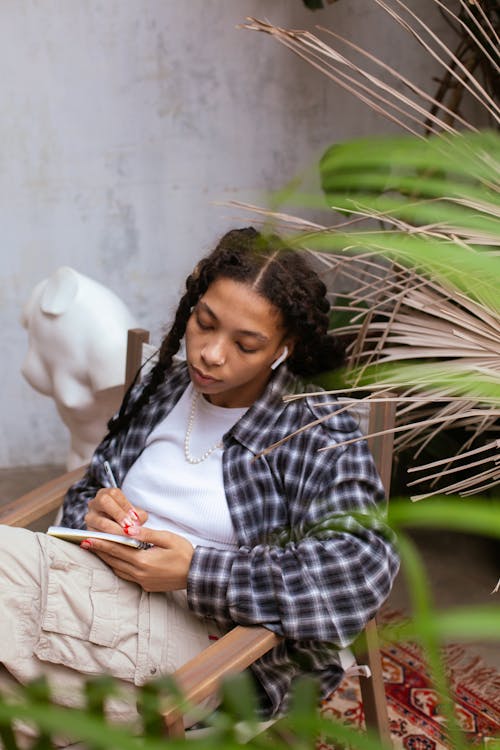 Woman in Plaid Shirt Writing on Notebook while Listening to Music