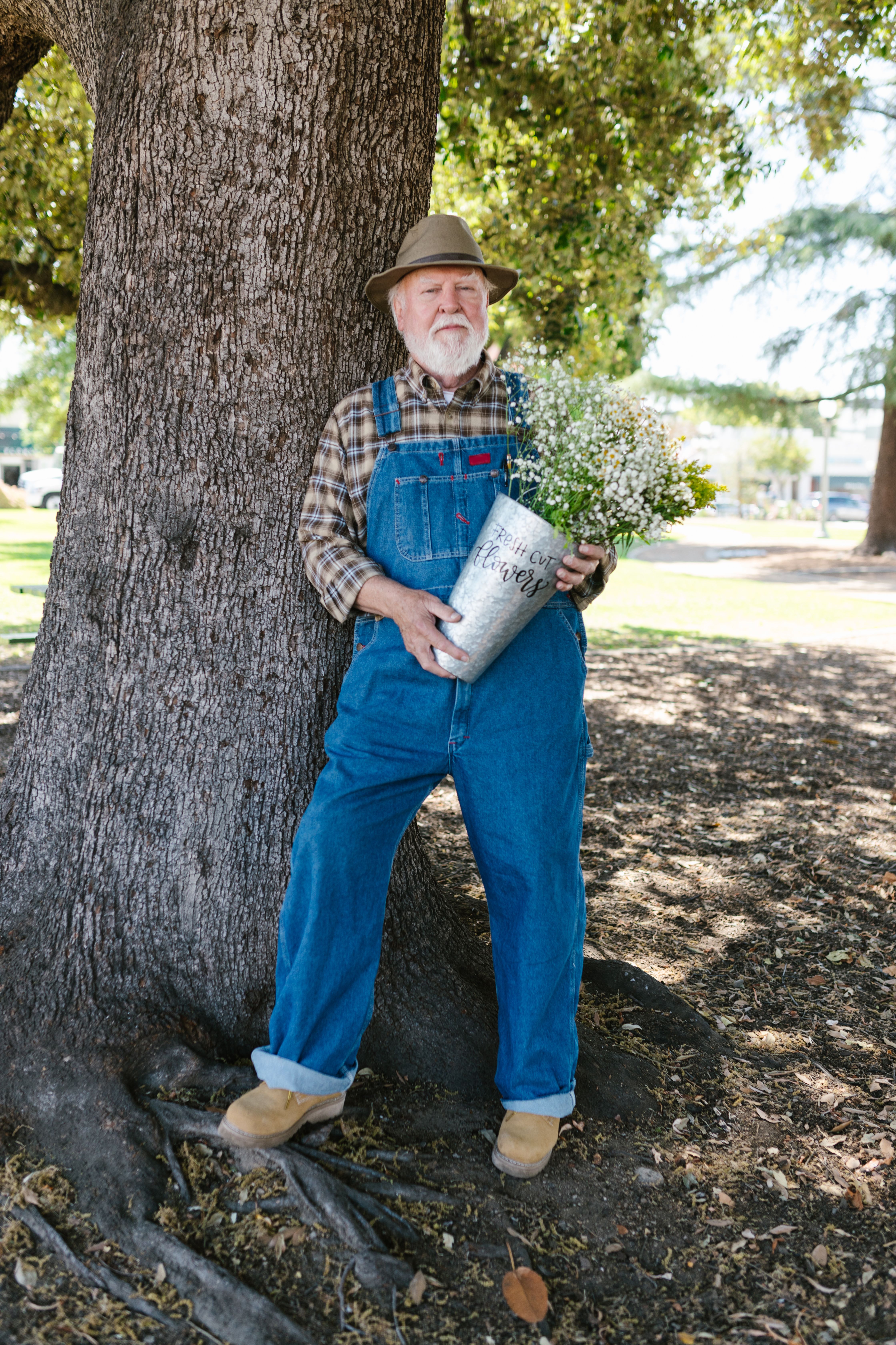 Senior man in overalls holding a flower bucket by a tree in a sunny park.