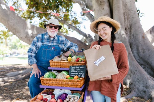 A Woman in Knitted Sweater Holding Paper Bag Near the Man Standing Near the Cart with Woven Baskets