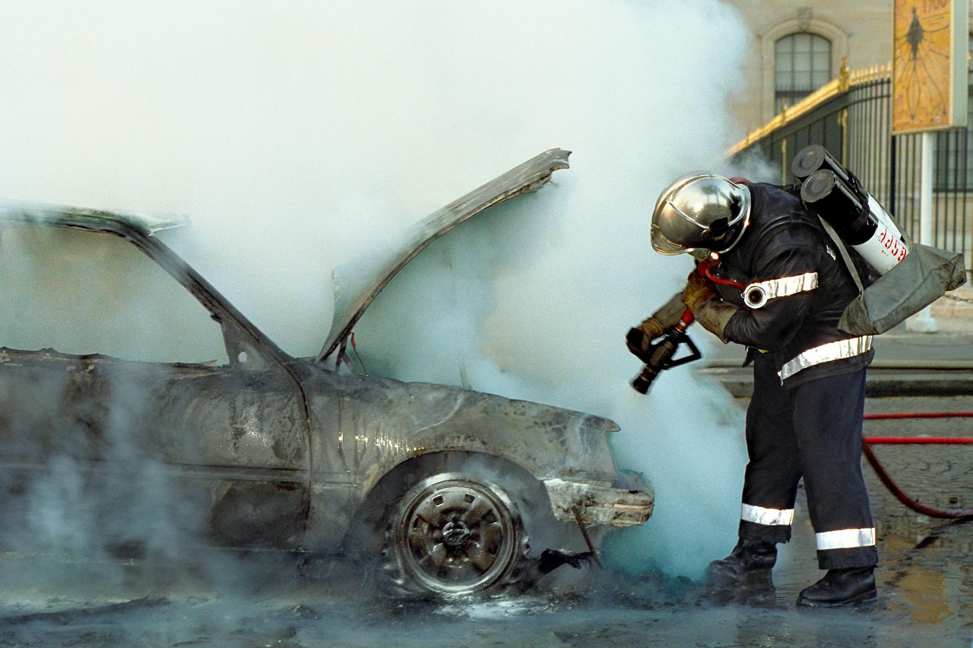 Firefighter using extinguisher on a smoking, damaged car in an urban area. High action scene.