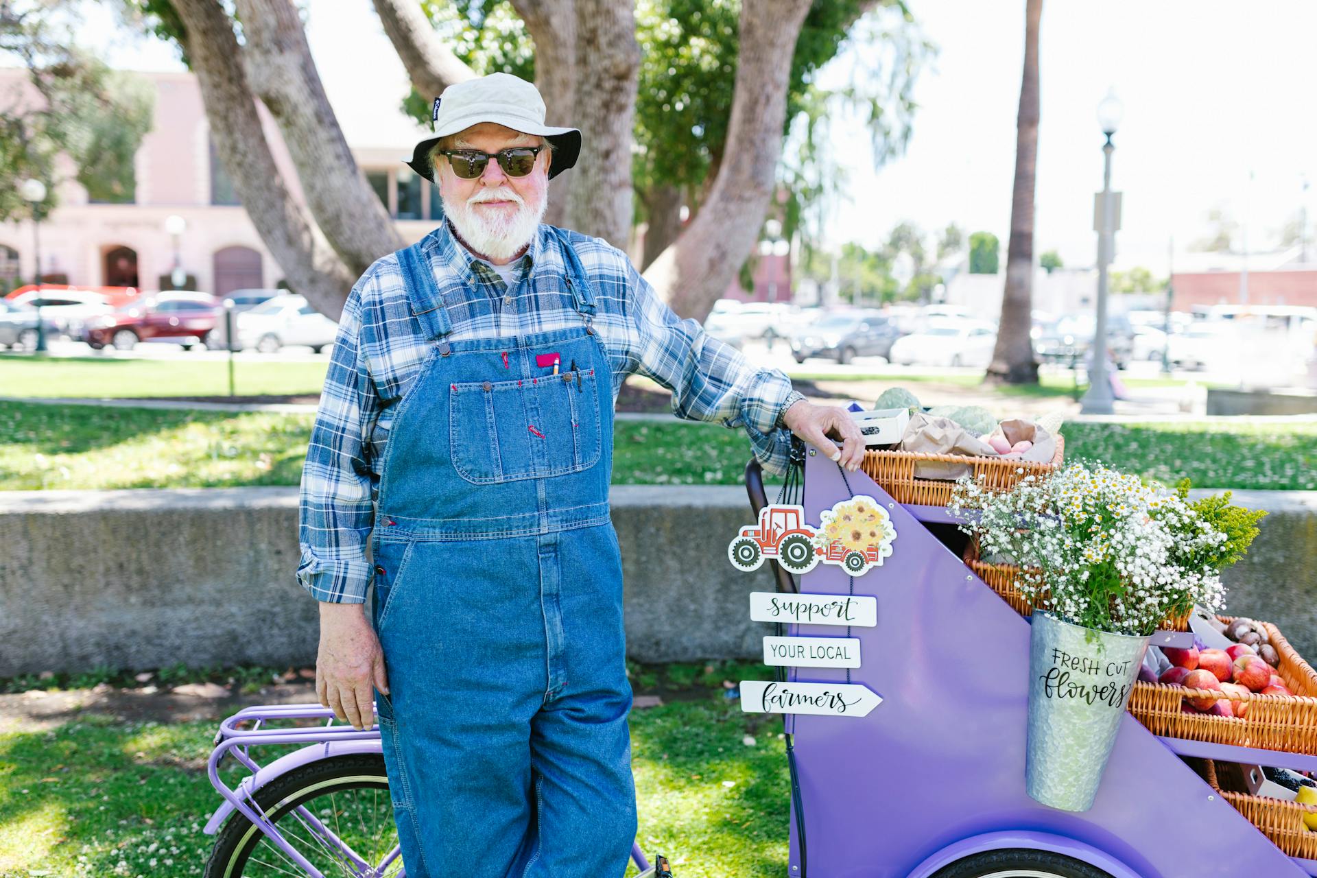 An elderly man selling fresh produce and flowers at an outdoor market. Support local farmers signage.