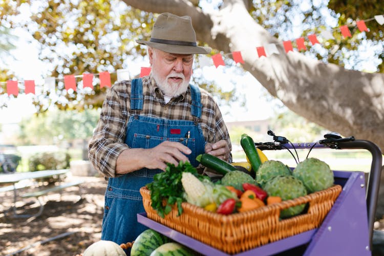 An Elderly Man Wearing Denim Overalls And A Hat Looking At Vegetables