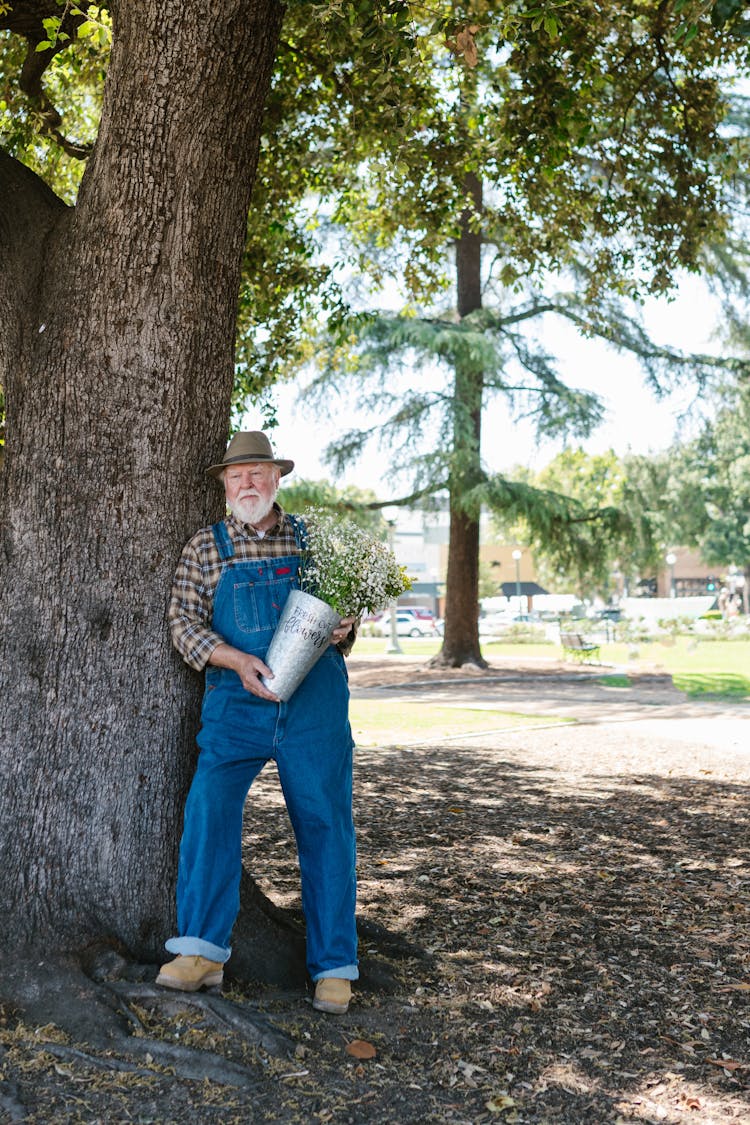 An Elderly Man Wearing Denim Overalls And A Hat Holding A Vase Of Flowers