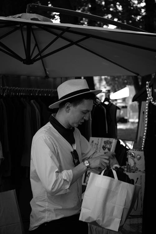 A Grayscale Photo of a Man Wearing a Hat while Holding a Paper Bag