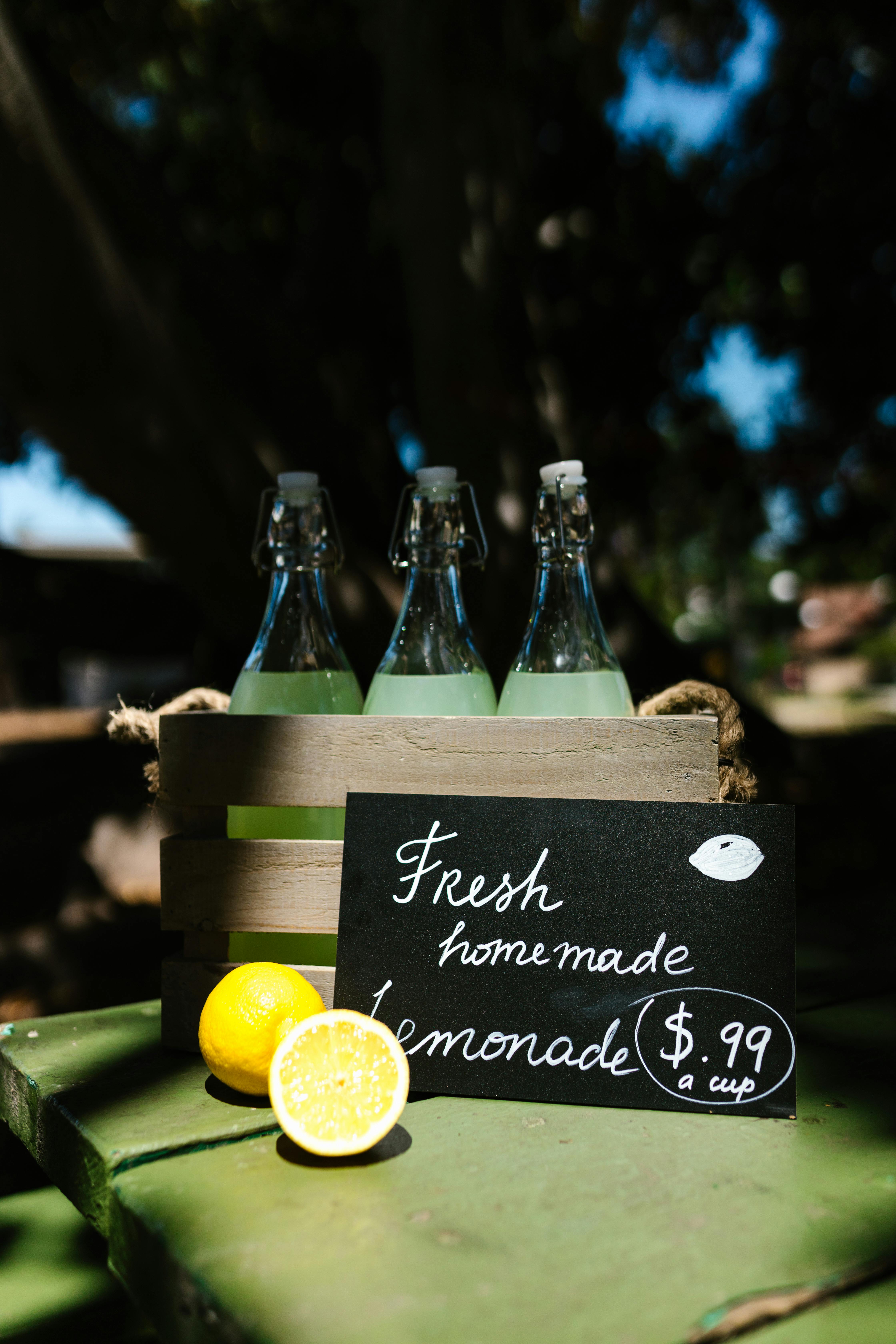 Fresh homemade lemonade bottles with a price sign, set outdoors in a sunny environment.