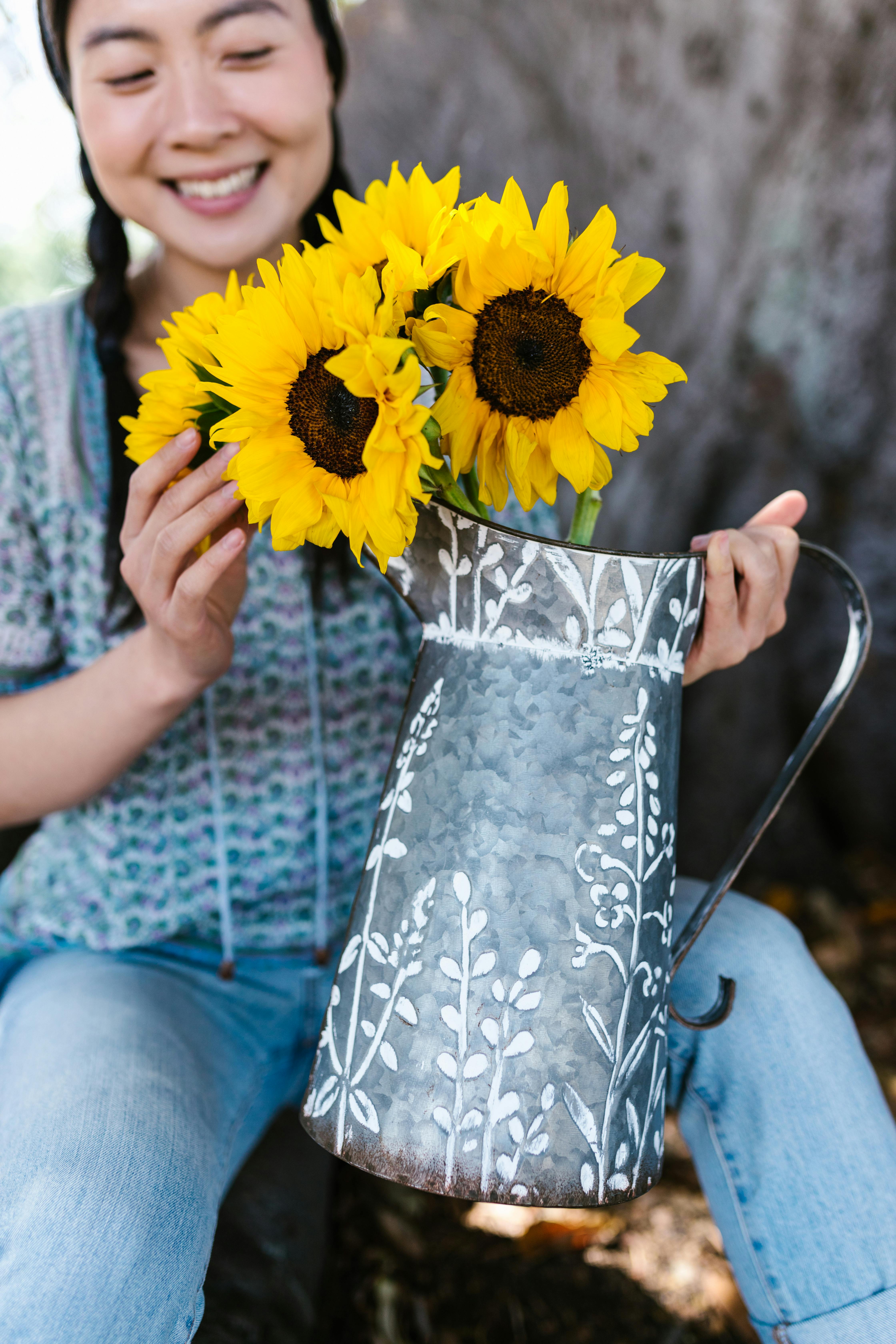 A woman holding a decorative metal pitcher filled with bright yellow sunflowers, exuding a cheerful mood.