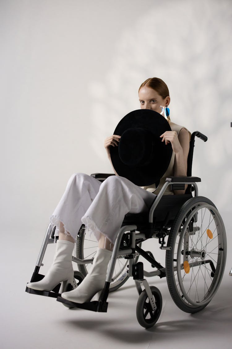 A Woman In White Pants Sitting On The Wheelchair While Holding A Black Hat