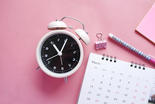 Close-Up Shot of an Alarm Clock and a Calendar on a Pink Surface