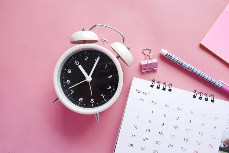 Close-Up Shot Of An Alarm Clock And A Calendar On A Pink Surface