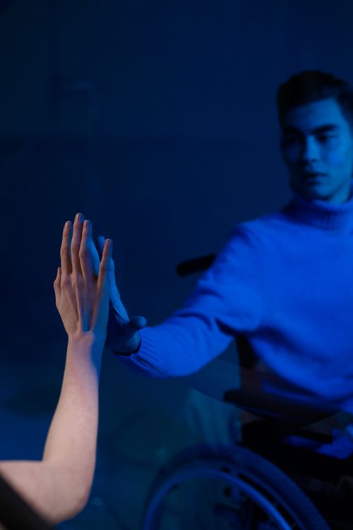 Free A man in a wheelchair reaching out to touch a hand in a blue-lit room, evoking connection. Stock Photo