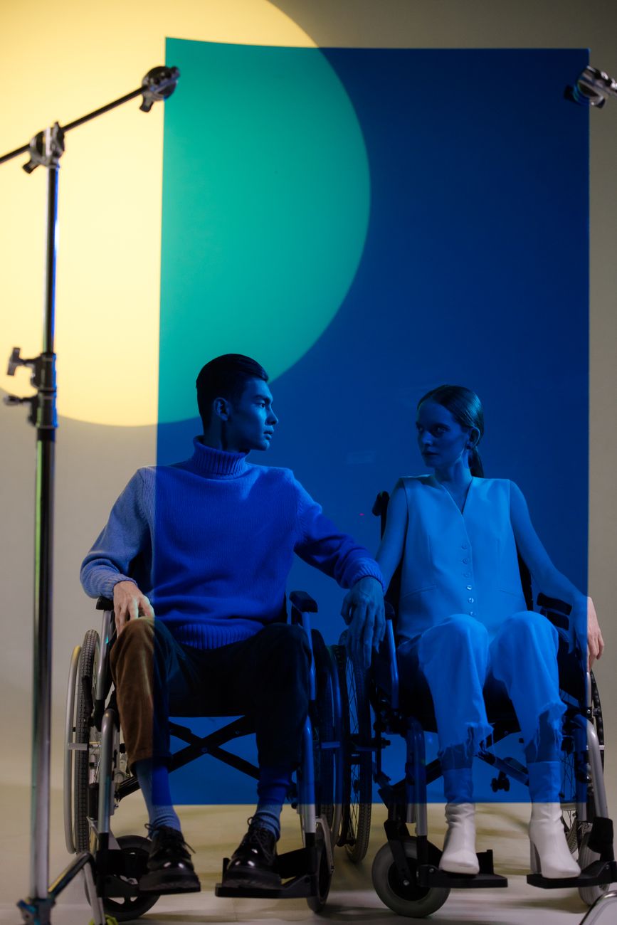 Un homme et une femme en fauteuil roulant, éclairés par des nuances de bleu et de jaune, se regardent dans un studio photo avec un fond coloré et des projecteurs professionnels.