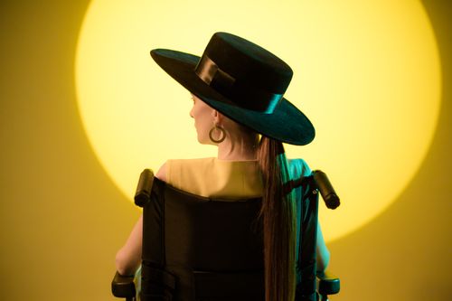 Free A woman in a wheelchair, wearing a stylish hat, is seated against a vibrant yellow backdrop. Stock Photo