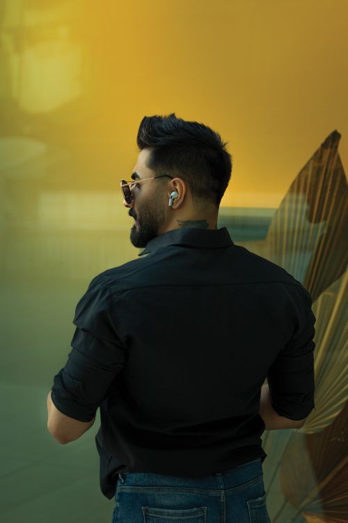 Free A fashionable man with sunglasses and earbuds, viewed from behind against a warm backdrop. Stock Photo