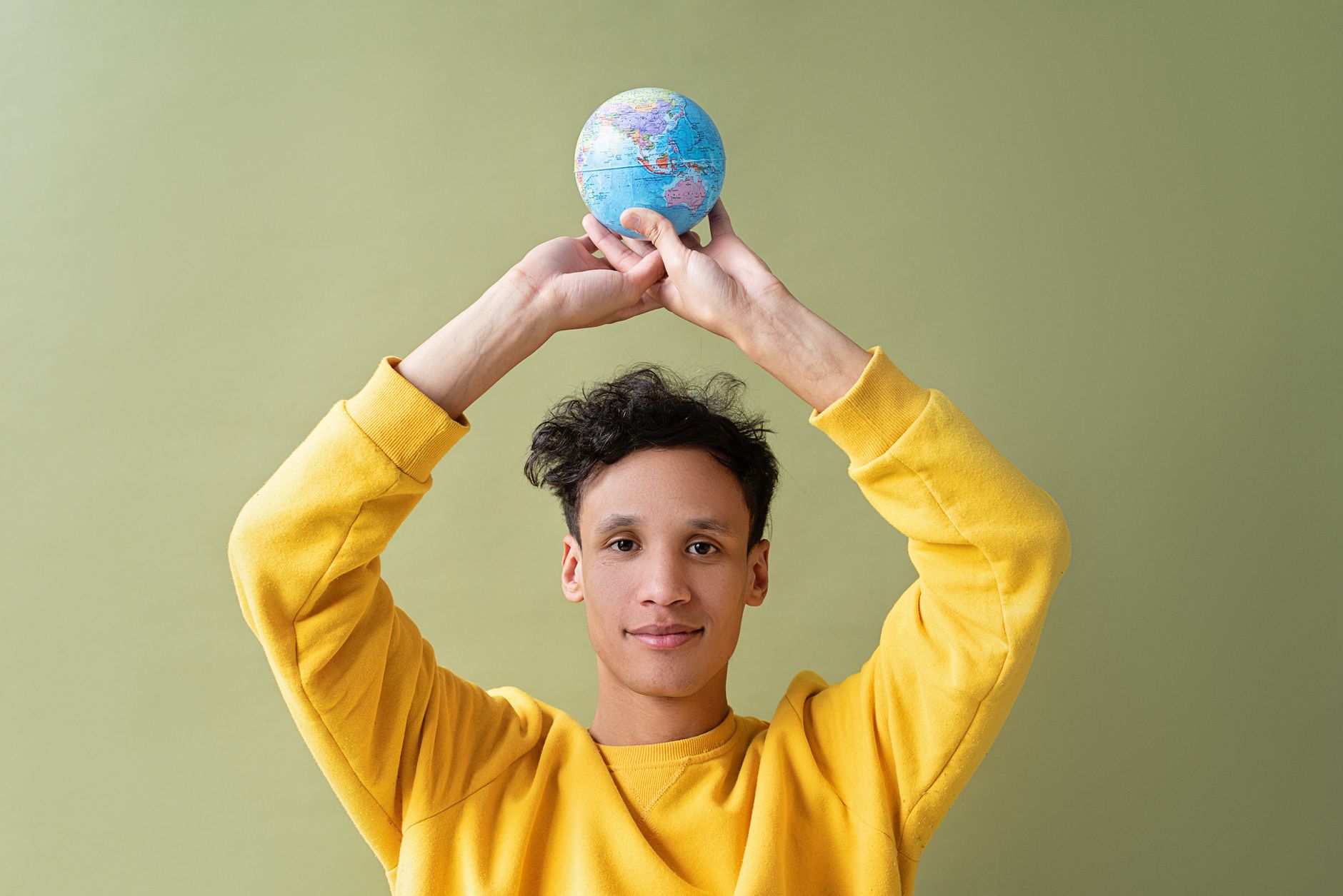 A young man in a yellow sweatshirt holds a globe above his head against a green background.