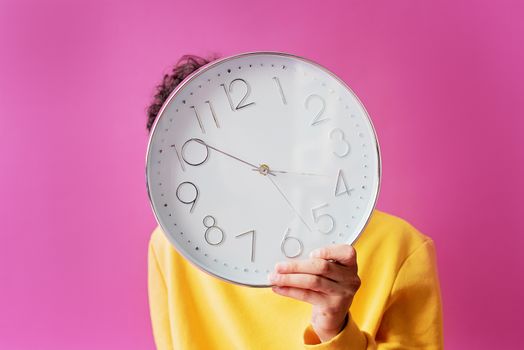 A person in a yellow shirt holds a large wall clock against a vibrant pink background.
